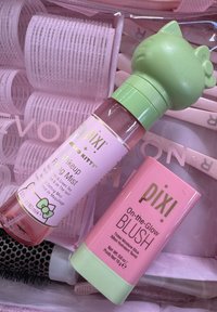 Produits cosmétiques roses et verts : un flacon vaporisateur avec un capuchon Hello Kitty et un bâton de blush rose. L'arrière-plan comprend des rouleaux à cheveux roses.