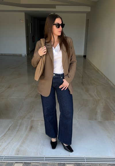 Mujer de pie en interiores con gafas de sol, blazer marrón, camiseta blanca, jeans oscuros, zapatos negros y llevando un bolso beige con textura colgado de un hombro.