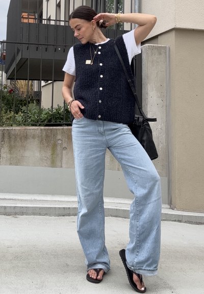 Mujer de pie al aire libre con un chaleco negro abotonado sobre una camiseta blanca, jeans de pierna ancha de un azul claro, sandalias negras, joyas doradas y un bolso negro de hombro.