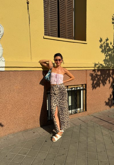 Modelo con un top sin tirantes rosa y una falda maxi de estampado de leopardo, usando gafas de sol, se apoya contra una pared amarilla. Sandalias y un bolso pequeño completan el look.