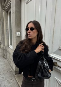 Musta lyhyt takki, tiukka musta paita, tumma housut, pyöreät mustat aurinkolasit ja musta nahkalaukku, jossa on hopeisia yksityiskohtia. Sileät pinnat ja selkeät linjat.