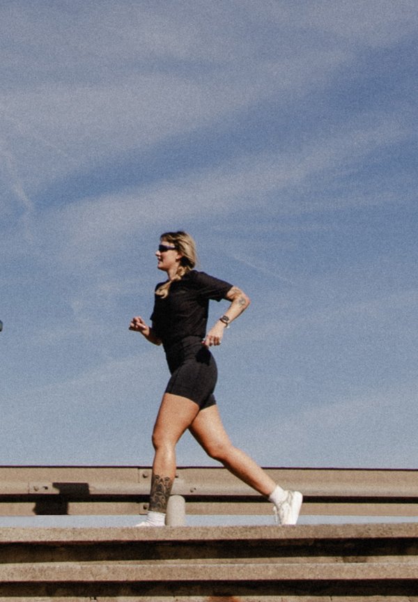 Una mujer vestida con ropa deportiva negra y zapatillas blancas corre por una pista al aire libre bajo un cielo despejado.