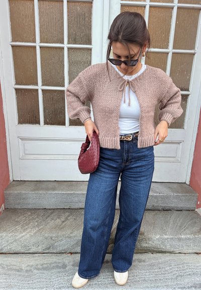 Cárdigan rosa claro de punto con mangas anchas, atado al cuello, llevado encima de una camiseta blanca. Vaqueros azules de pierna ancha y bolso trenzado burdeos.