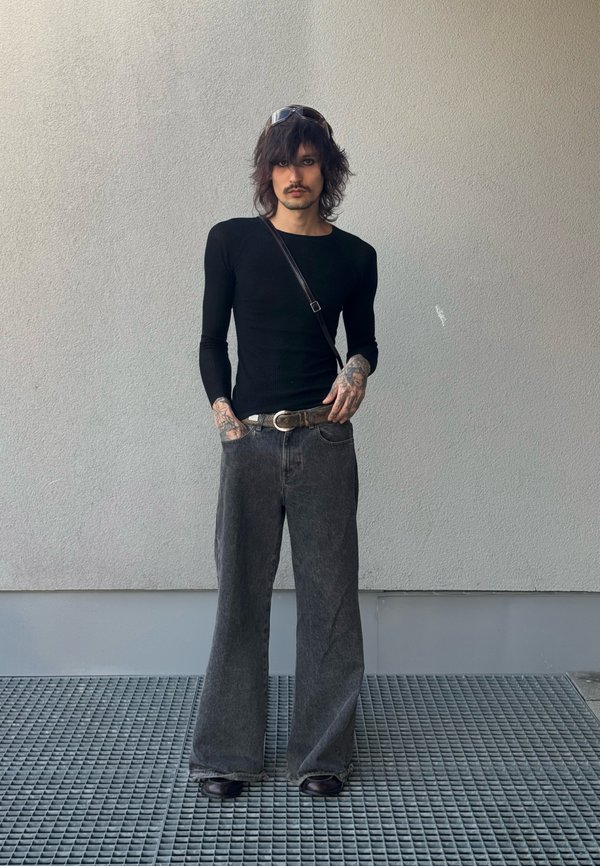 Personne mince avec des tatouages portant une chemise noire à manches longues, un jean gris large, des chaussures noires et des lunettes de soleil sur la tête, debout contre un mur gris.