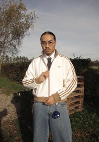 Casaco desportivo branco com riscas castanhas, combinado com jeans azuis largos. Acessórios incluem óculos e um porta-chaves azul. Ambiente exterior.