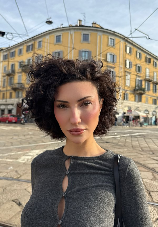 Haut en tricot gris avec un col rond et des détails de boutons décoratifs. Texture douce, coupe près du corps. Sac à main noir visible. Fond urbain avec un bâtiment jaune.