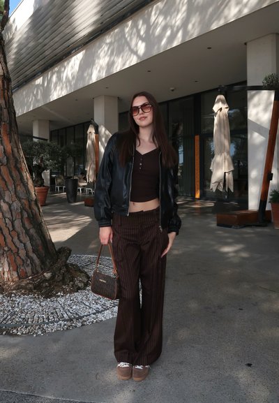 Joven mujer con gafas de sol, chaqueta de cuero negra, pantalones marrones a rayas y zapatillas está de pie al aire libre cerca de un árbol y la entrada de un edificio moderno.