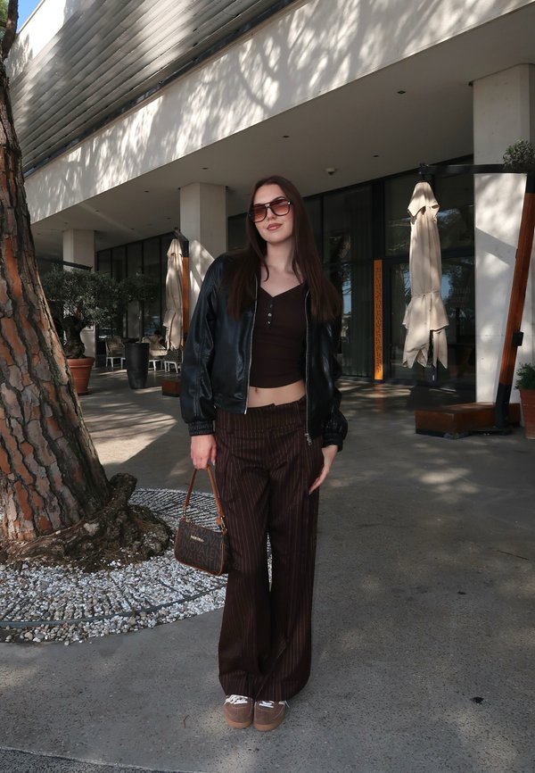 Joven mujer con gafas de sol, chaqueta de cuero negra, pantalones marrones a rayas y zapatillas está de pie al aire libre cerca de un árbol y la entrada de un edificio moderno.