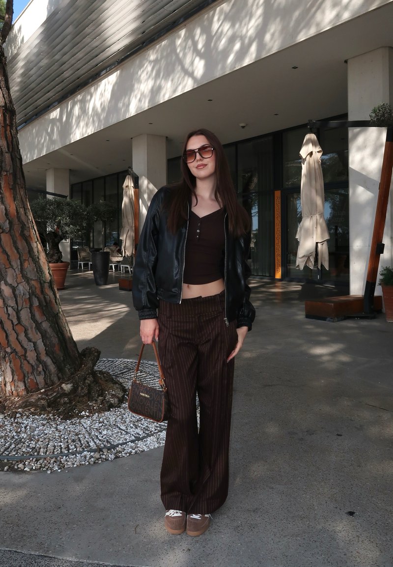 Jeune femme portant des lunettes de soleil, une veste en cuir noire, un pantalon rayé marron et des baskets, se tient dehors près d'un arbre et de l'entrée d'un bâtiment moderne.