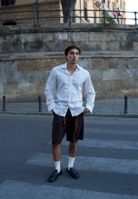 Balta ilgomis rankovėmis marškiniai, rudi šortai su diržais, balti kojinės, juodi batai. Asmuo stovi gatvėje su tekstūruota akmens siena fone.