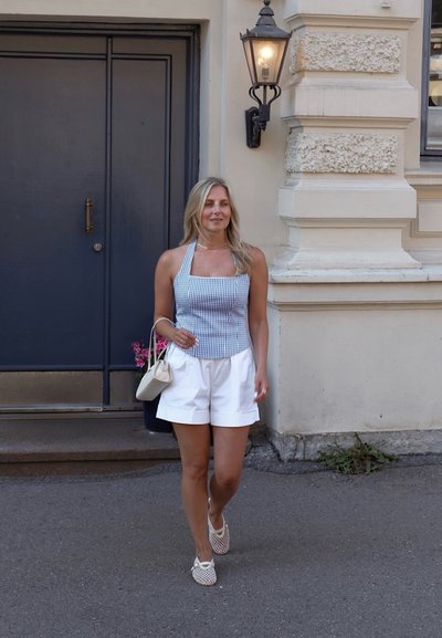 Top sin mangas a cuadros azul y blanco, pantalones cortos blancos ajustados y bolso blanco. Una mujer con zapatos blancos de tiras pasa junto a una puerta.