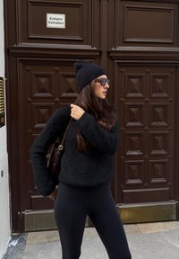 Črn puhast pulover, črne legice in črna kapa, dopolnjeni s temnimi sončnimi očali in rjavo torbico čez ramo, na rjavih vratih.