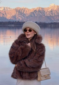 Femme en manteau de fourrure marron, chapeau beige et lunettes de soleil, debout au bord d'un lac calme avec des montagnes enneigées illuminées par le coucher du soleil en arrière-plan.
