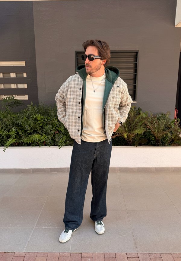 Muž s brýlemi, v kostkované bundě přes zelenou kapuci, bílém tričku, tmavých širokých kalhotách a bílých teniskách stojí venku s rukama v kapsách.