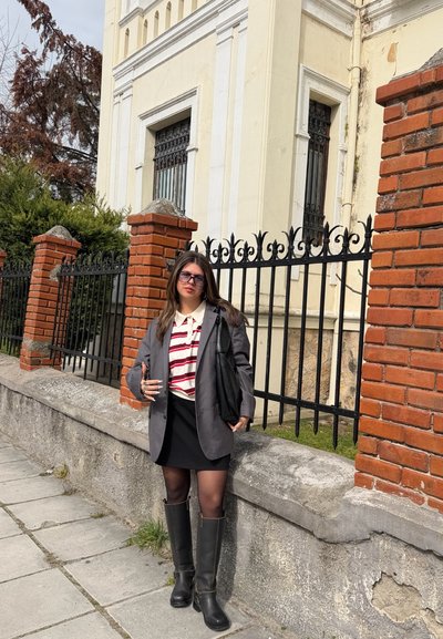 Joven mujer con blazer gris, camisa a rayas, falda negra y botas, de pie junto a una valla de ladrillo y hierro cerca de un edificio de color claro.