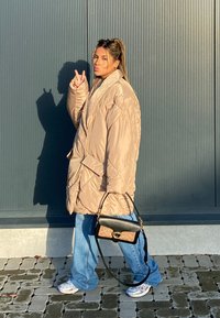 Bežani pufast kaput s širokim rukavima, plavi široki traperice i crna torbica. Srebrne tenisice upotpunjuju odjevnu kombinaciju. Pozadina prikazuje sivi zid s uspravnim rebrima.