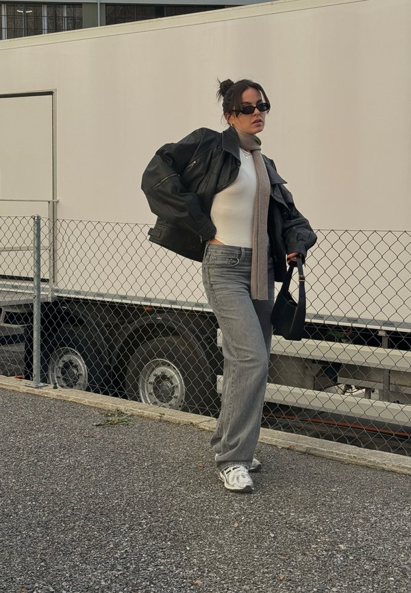 Chaqueta de cuero negra sobre un suéter blanco de cuello alto, jeans grises y zapatillas grises. Notables gafas de sol negras y una bufanda marrón clara, junto con un bolso.