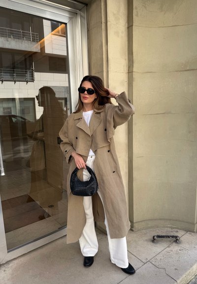 Mujer con gabardina beige, pantalones blancos, zapatos negros y gafas de sol, sosteniendo un bolso negro tejido, apoyada junto a una puerta de cristal y una pared de piedra.