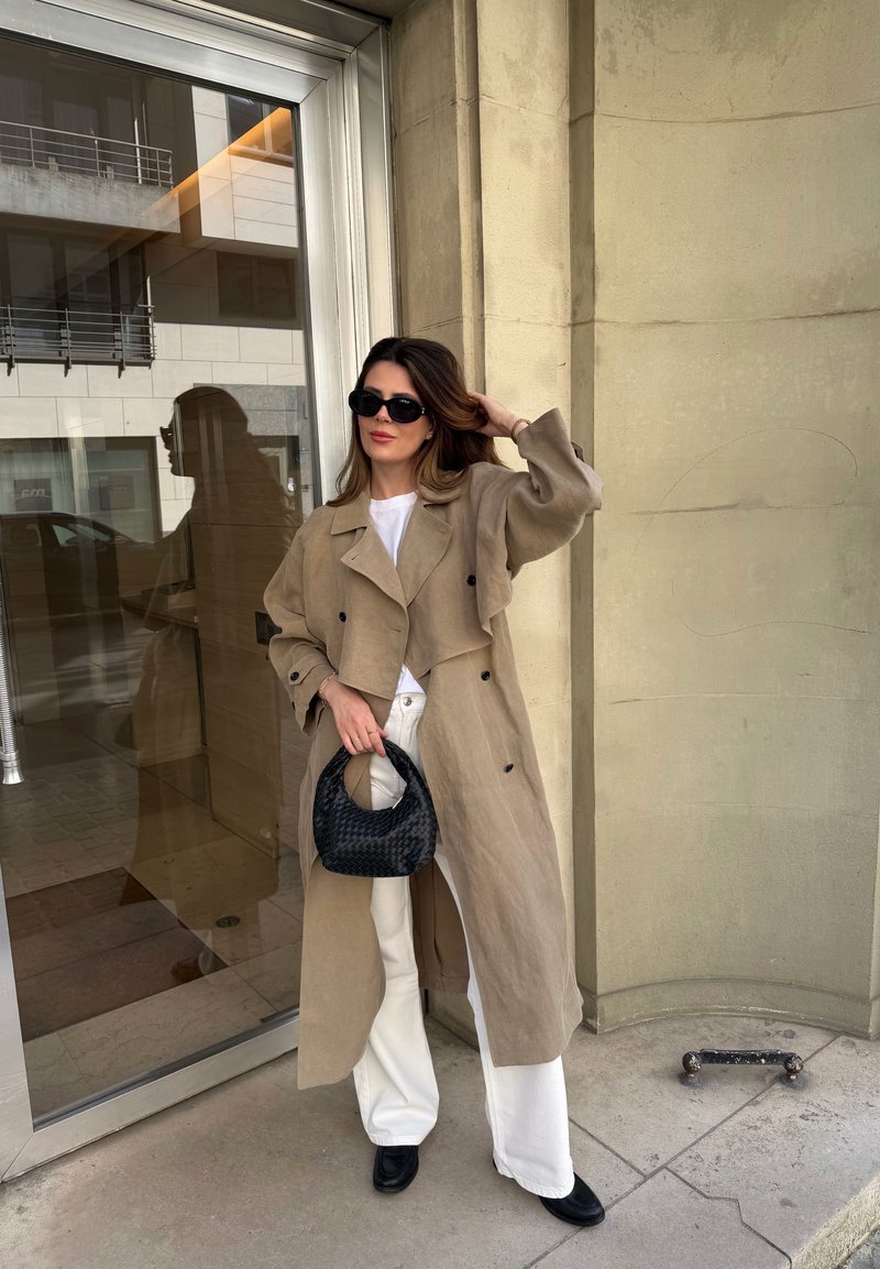 Donna con trench beige, pantaloni bianchi, scarpe nere e occhiali da sole, che tiene una borsa nera intrecciata, in piedi accanto a una porta di vetro e un muro di pietra.