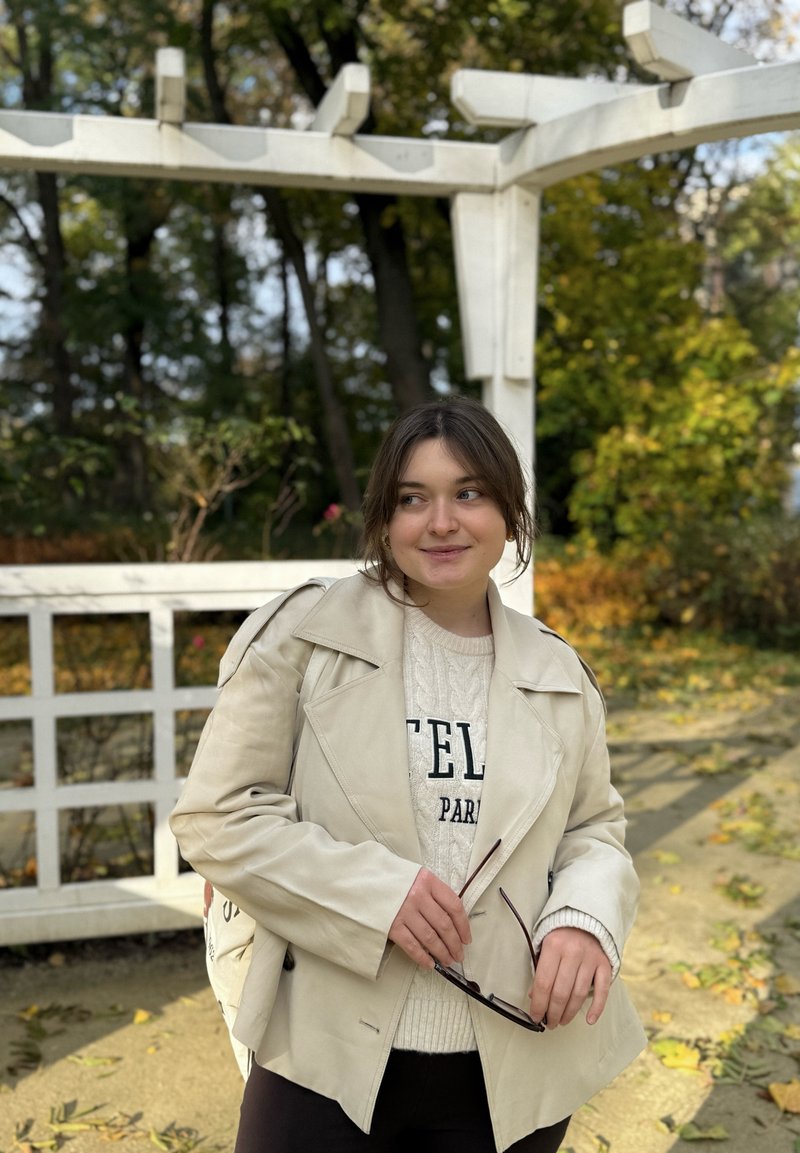 Veste en cuir beige clair avec un large col, portée sur un pull crème à torsades, associée à un pantalon foncé et tenant des lunettes de soleil.