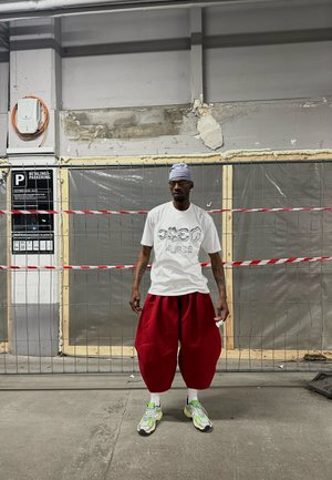 Camiseta blanca con logo en relieve, pantalones rojos de gran tamaño y zapatillas deportivas multicolores. El fondo presenta elementos de construcción y barreras.