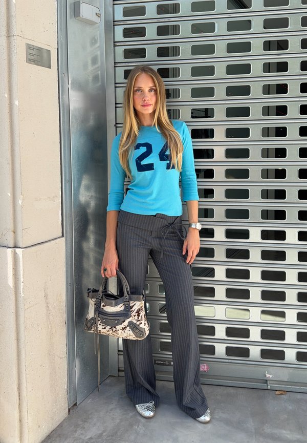 Jeune femme portant un haut bleu avec le numéro 24, un pantalon gris à fines rayures, des chaussures argentées, tenant un sac à main à motifs, debout près d'une porte métallique à volet roulant.