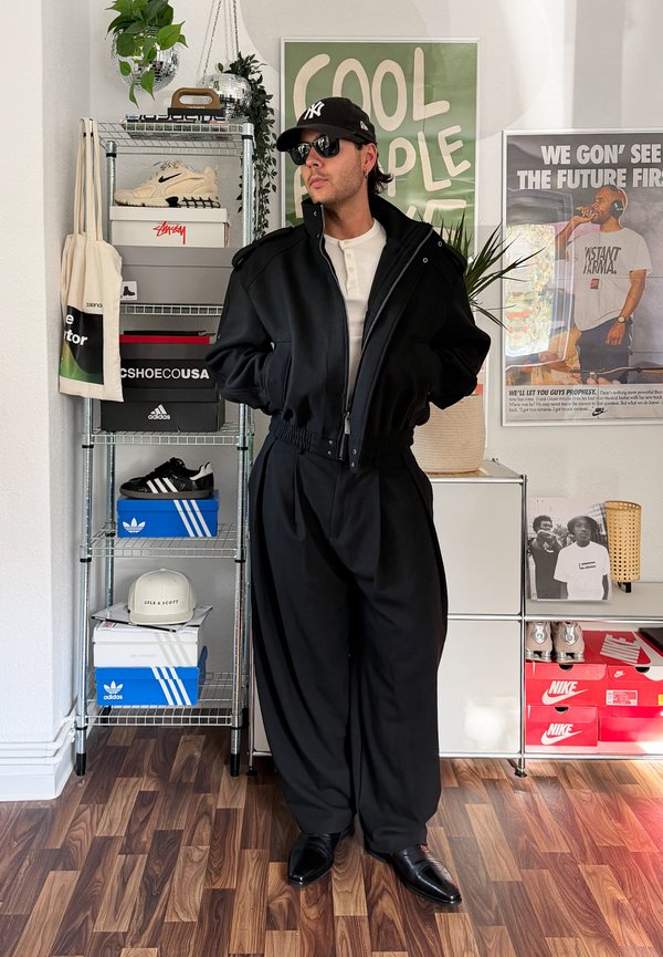Hombre con chaqueta negra, pantalones negros anchos, gafas de sol y gorra de béisbol, de pie en una habitación con cajas de zapatos y carteles en la pared.
