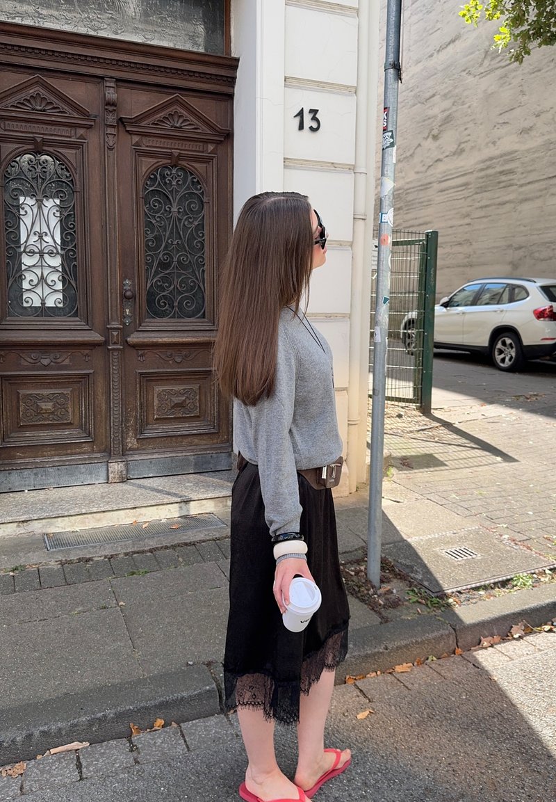Kvinne som står på siden, iført en grå genser, svart skjørt med blonder, holder en kaffekopp, sandaler, med langt hår og solbriller.
