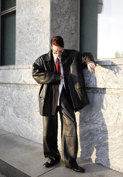 Chaqueta grande de cuero negro con bolsillos grandes, camisa blanca y corbata roja, combinada con pantalones anchos de cuero negro y zapatos negros.