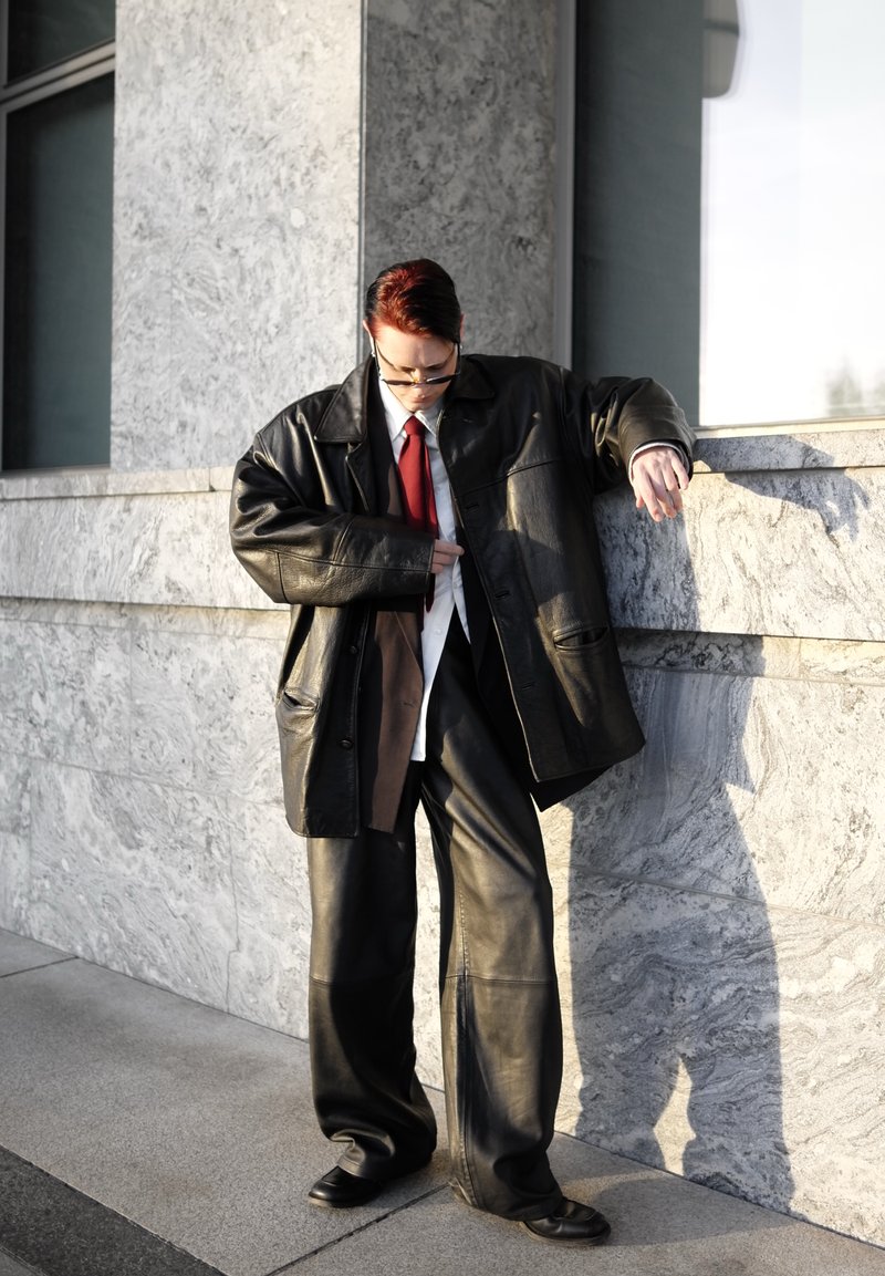 Personne aux cheveux rouges portant une grande veste en cuir noir et un pantalon, une chemise blanche et une cravate rouge, ajustant sa veste tout en s'appuyant sur un mur en pierre.