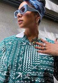 Camisa verde con estampado y diseños blancos, gafas de sol azules, collares de oro en capas y anillos multicolores. Tejido texturizado y accesorios distintivos.