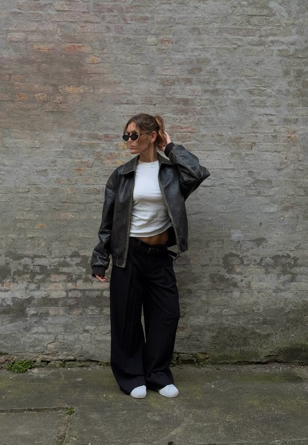 Une femme portant des lunettes de soleil, une veste en cuir noire, un haut blanc et un pantalon noir ample pose devant un mur en pierre.