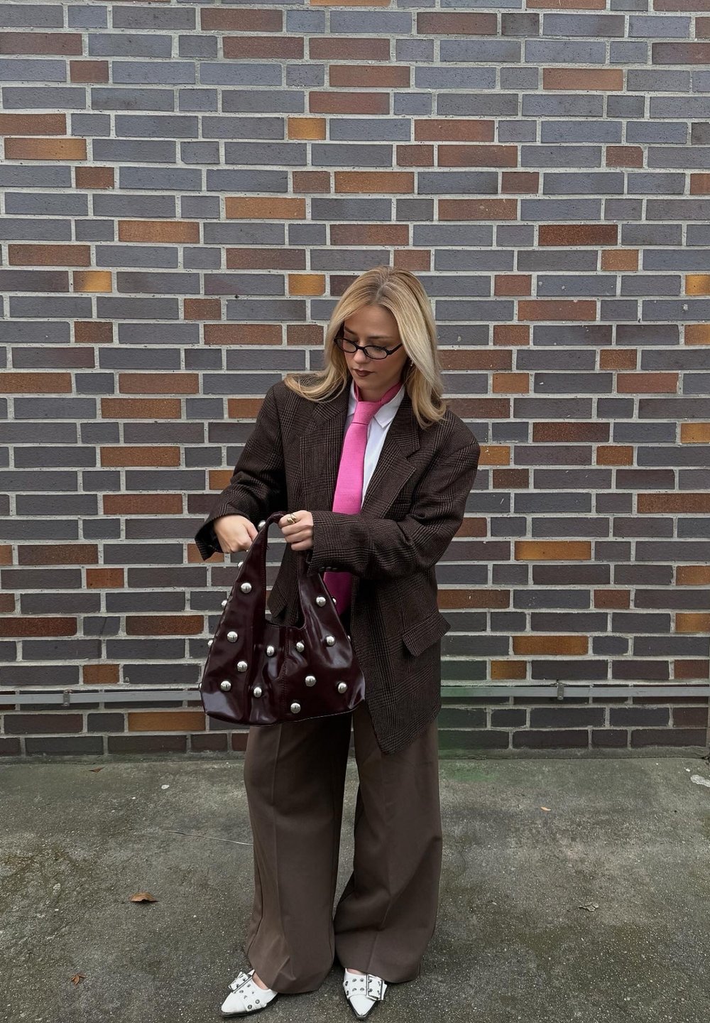 Hnedé kárované oversize sako, biela košeľa, ružová kravata, hnedé široké nohavice, drží lesklú vínovočervenú kabelku so striebornými cvočkami.