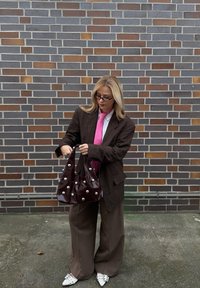 Hnedé kárované oversize sako, biela košeľa, ružová kravata, hnedé široké nohavice, drží lesklú vínovočervenú kabelku so striebornými cvočkami.