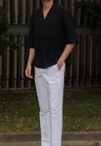 Čierno-biele pruhované krátke tričko s voľným strihom a gombíkmi, v kombinácii s bielymi rovnými nohavicami. Pozadie tvorí drevené panely a tráva.