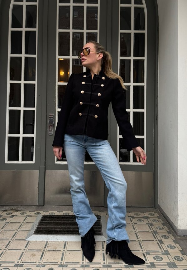 Kvinde med hestehale iført sort militærinspireret jakke, solbriller, lyseblå vidde jeans og sorte støvler med frynser, stående ved en dør med glaspaneler.