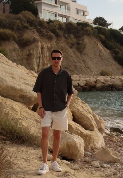 Figura masculina con una camisa oscura de manga corta y botones, pantalones cortos blancos y zapatillas blancas, de pie en una playa rocosa con acantilados detrás.