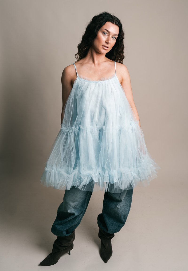 Nő hosszú, sötét, göndör hajjal, áttetsző világoskék ujjatlan ruhát visel laza farmernadrág fölött, fekete hegyes orrú csizmával, egyszerű háttér előtt.