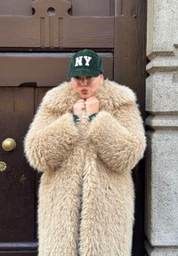 Cappotto di pelliccia beige e morbida con maniche oversize, indossato sopra una maglia verde. Berretto verde scuro con "NY" in bianco. Orecchini a cerchio dorati.