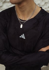 Oseba, ki nosi tri srebrne verižice ter črno majico Adidas, z crossed arms (prečkanimi rokami) proti teksturirani ozadju.