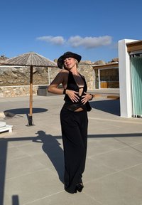 Črni top z naramnicami in odprtimi hrbtnimi deli, v kombinaciji z ohlapnimi črnimi hlačami. Dodatki vključujejo širokokrajni črni klobuk. Zunanje okolje s kamnito ozadje.