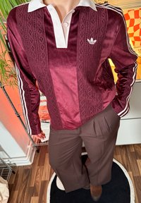 Burgunder rød langermet satengskjorte med hvit krage, intrikat blondermønster og hvite striper på ermene, kombinert med brune plissert bukser.