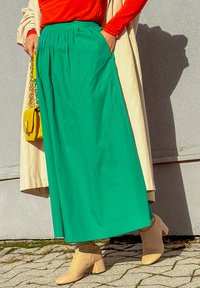 Grüner, fließender Maxirock mit Falten, kombiniert mit einem figurbetonten roten Oberteil. Ergänzt durch eine gelbe Handtasche und beigefarbene Stiefeletten.