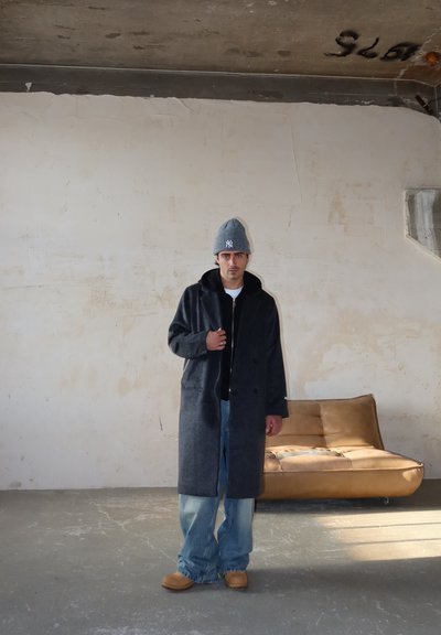 Hombre con gorro gris, abrigo largo oscuro, vaqueros holgados y zapatos marrones se encuentra en una habitación industrial junto a un sofá de cuero marrón.
