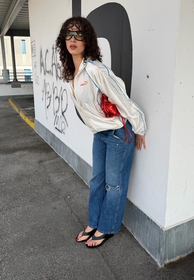Joven mujer con gafas de sol apoyada contra una pared blanca cubierta de grafitis, vistiendo una chaqueta blanca de Puma, vaqueros azules rotos, sandalias negras y sosteniendo un bolso rojo.