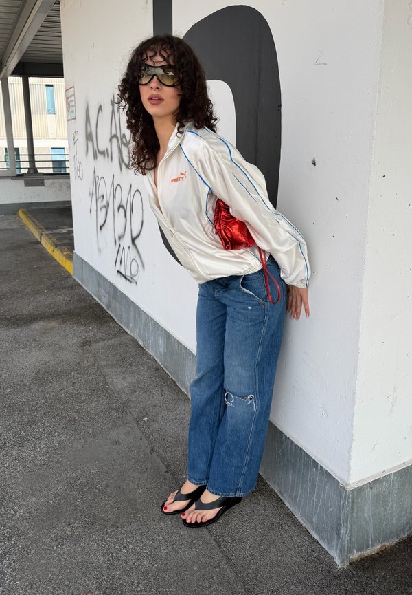 Ung kvinde i solbriller læner sig op ad en hvid væg dækket af graffiti iført en hvid Puma-jakke, revnede blå jeans, sorte sandaler og holder en rød taske.