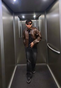 Jeune homme portant des lunettes de soleil, une veste en cuir marron et un jean noir, debout dans un ascenseur métallique avec une rampe et des luminaires au plafond.