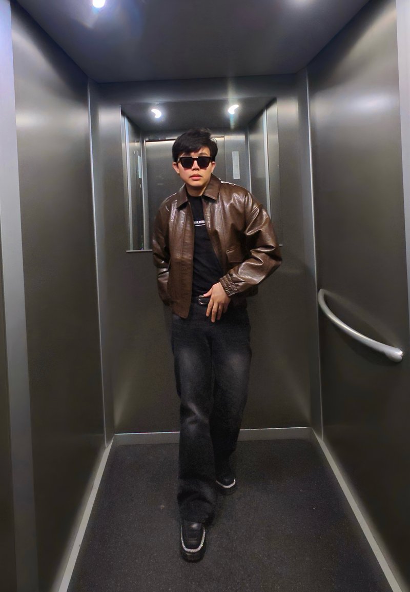 Jeune homme portant des lunettes de soleil, une veste en cuir marron et un jean noir, debout dans un ascenseur métallique avec une rampe et des luminaires au plafond.