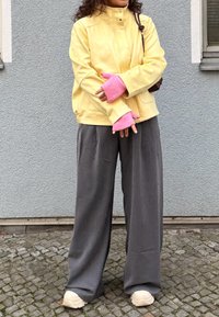 Giacca gialla in finta pelle scamosciata con colletto alto, guanti senza dita rosa a coste, pantaloni grigi a gamba larga e sneakers bianche su pavimentazione acciottolata.