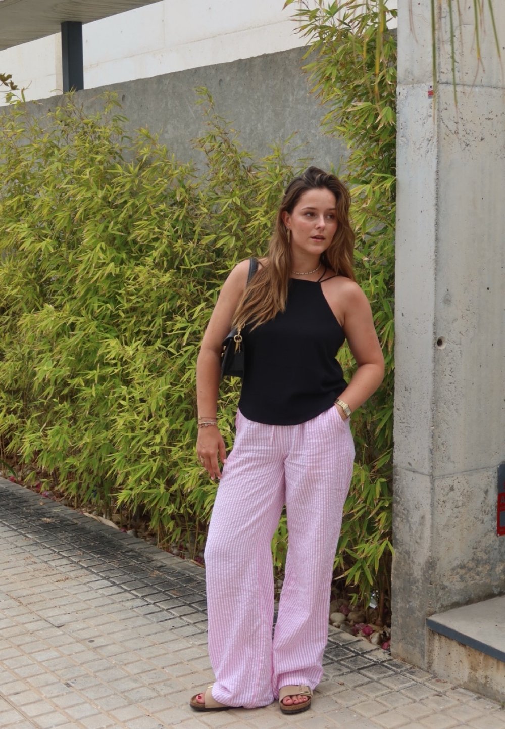 Μαύρη αμάνικη μπλούζα με ψηλό λαιμό, ροζ και άσπρες ριγέ παντελόνες φαρδιές, και καφέ σανδάλια, σε φόντο με πράσινα φυτά.